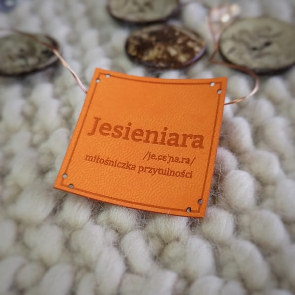 Pomarańczowa naszywka Jesieniara z ciemnym napisem, widoczna linia pomocnicza do szycia.