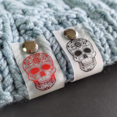 Dwie metki Calavera zamocowane na niebieskim ściągaczu swetra, pokazujące efekt przebijania koloru przez silikon.