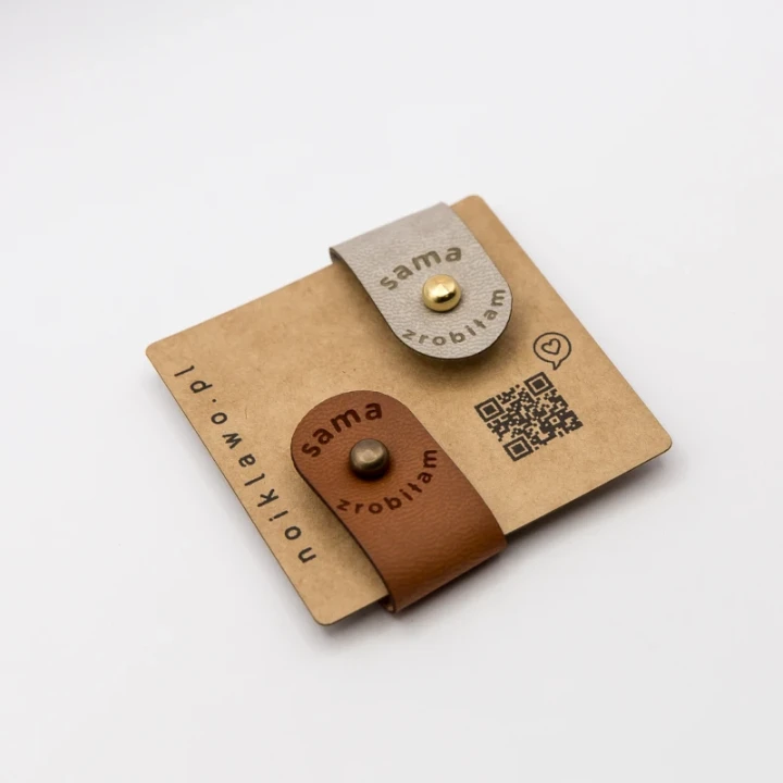 Dwie metki z ekoskóry z napisem Sama zrobiłam w kolorach brązowym i beżowym, zamocowane na skręcane nity do czapki.