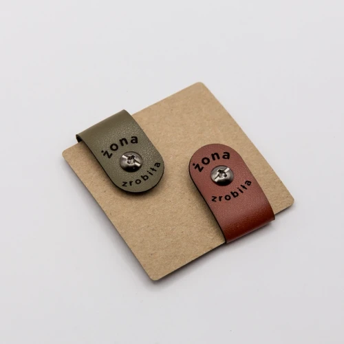 Metki w kolorach ziemi ceglastym i oliwkowym z napisem „Żona zrobiła” i czarnym nitem.